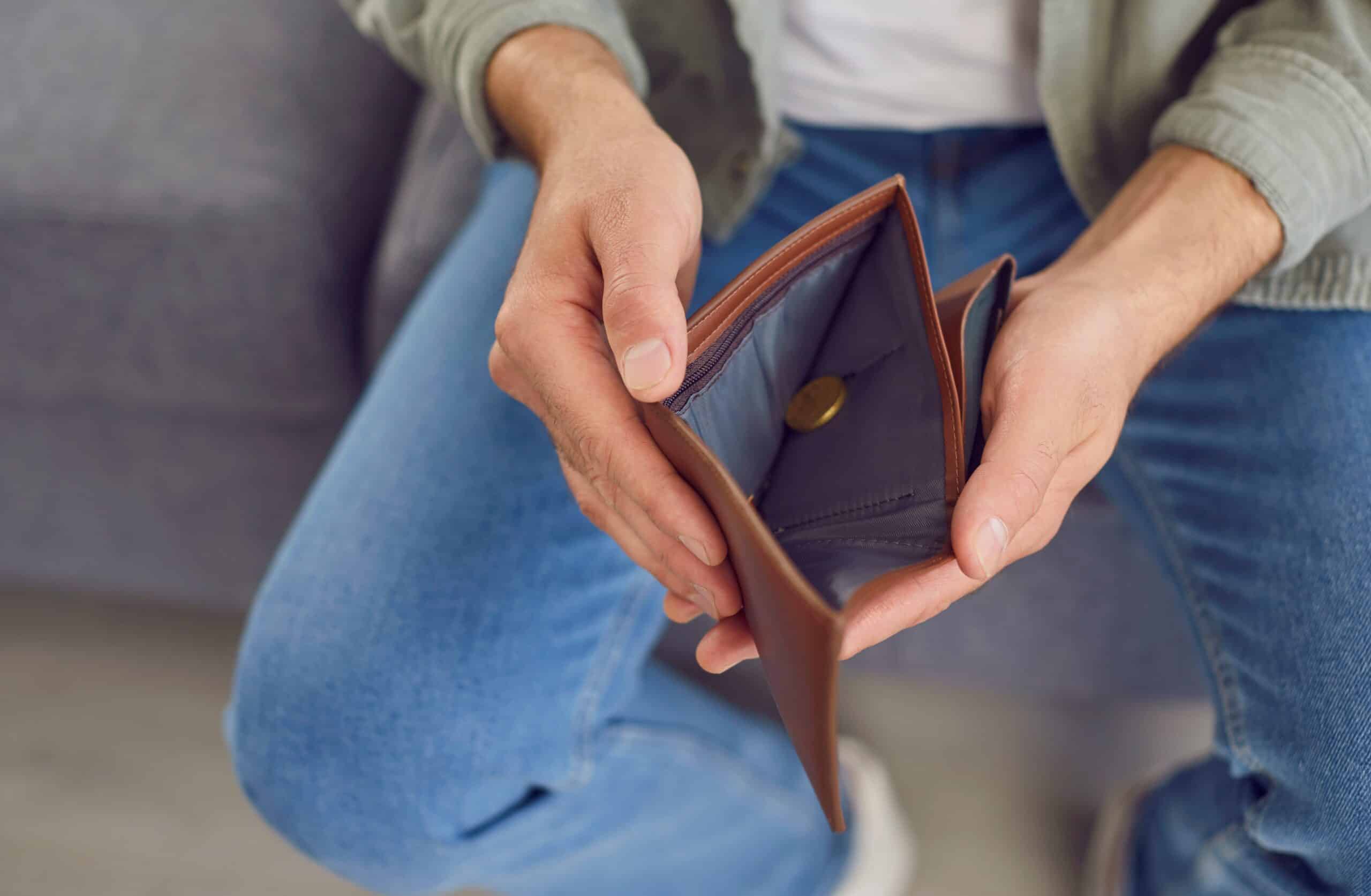 A person in jeans and a jacket sits holding an open wallet that appears mostly empty except for a single coin inside.