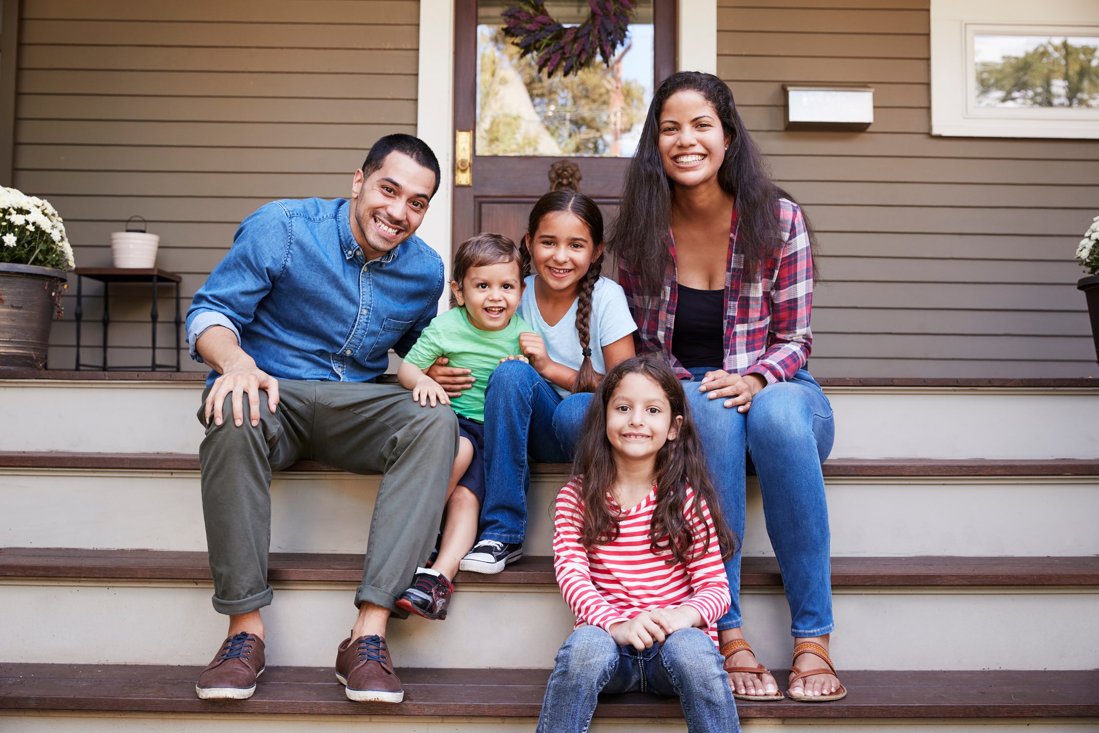 A smiling family of five sits on front steps, two adults and three kids enjoying a sunny day with door and potted plants behind them.