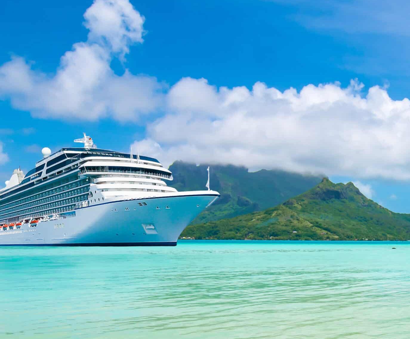 A large white cruise ship anchored in turquoise water near a tropical island with palm trees, green mountains, and blue sky.