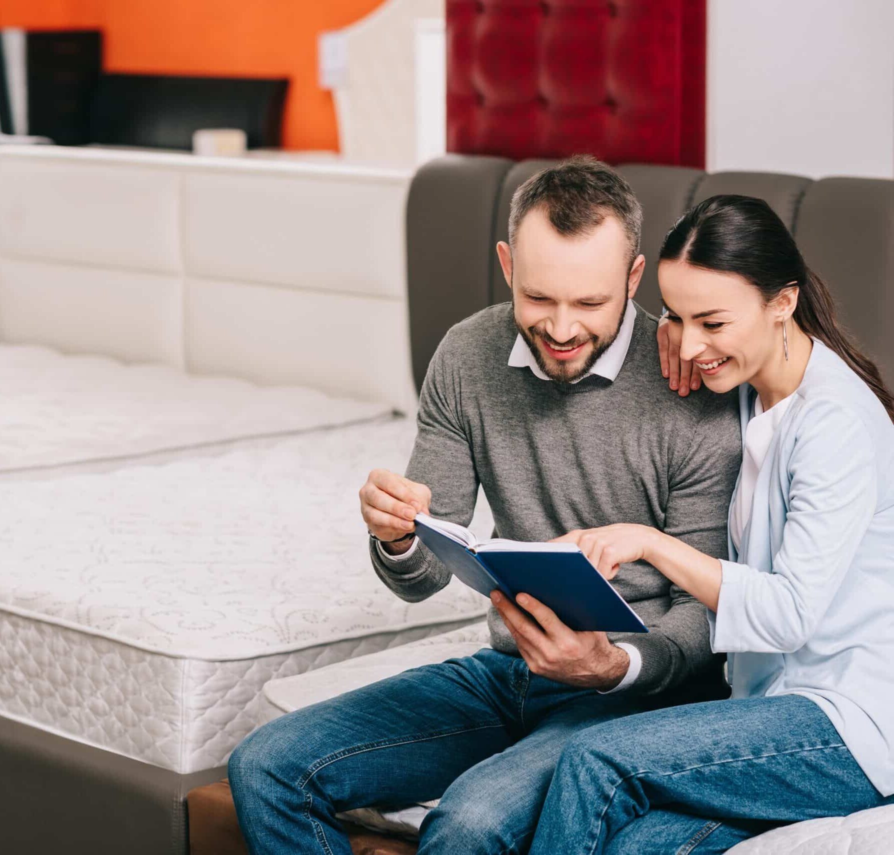 A happy couple sits together on a bed in a mattress store, reviewing a booklet, with various mattresses visible in the background.