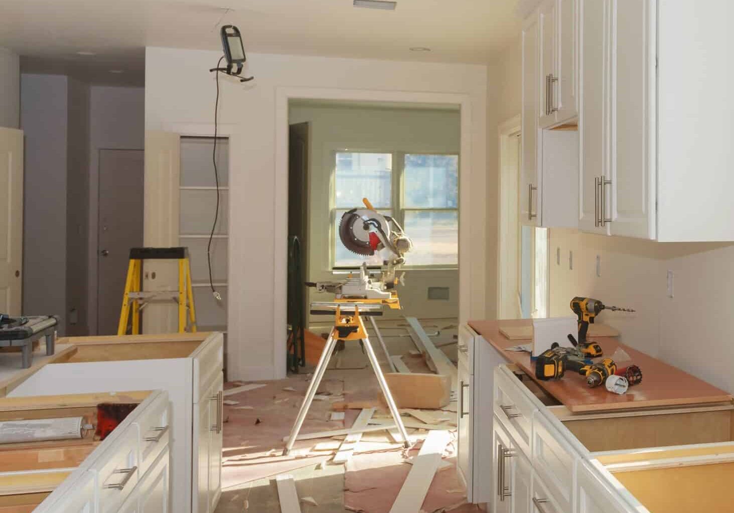 Kitchen renovation scene with white cabinets going in, tools scattered on counters, an unfinished island, and sunlight through windows.