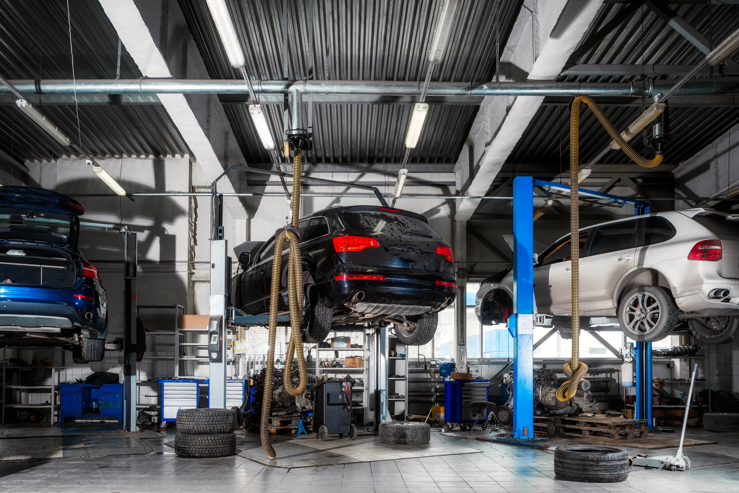 Modern auto repair shop with three cars on hydraulic lifts, scattered tools and spare tires, all brightly lit by overhead lights.