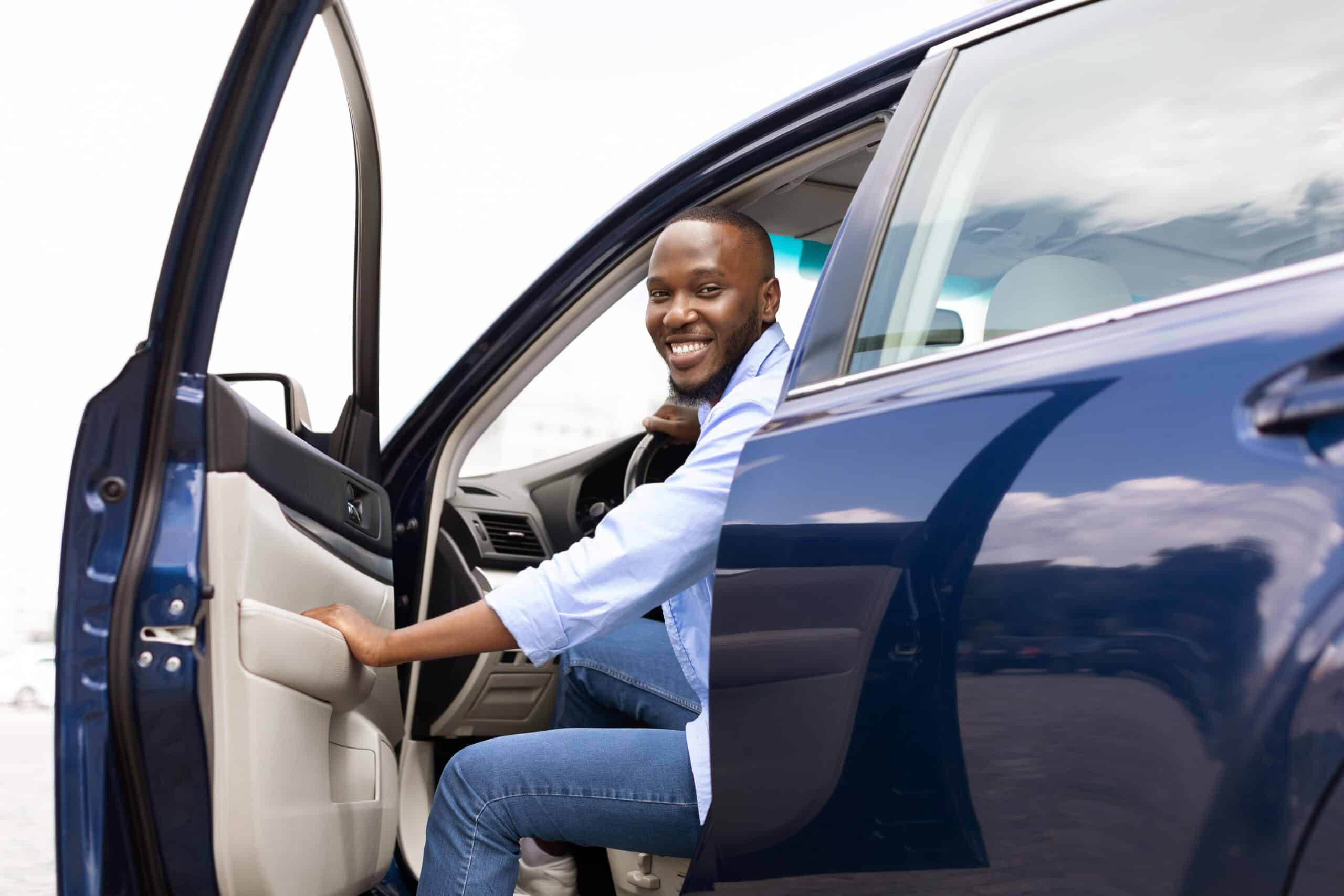 A smiling man sits in the driver’s seat of a blue car with the door open, looking at the camera. Sky and clouds reflect on the car.