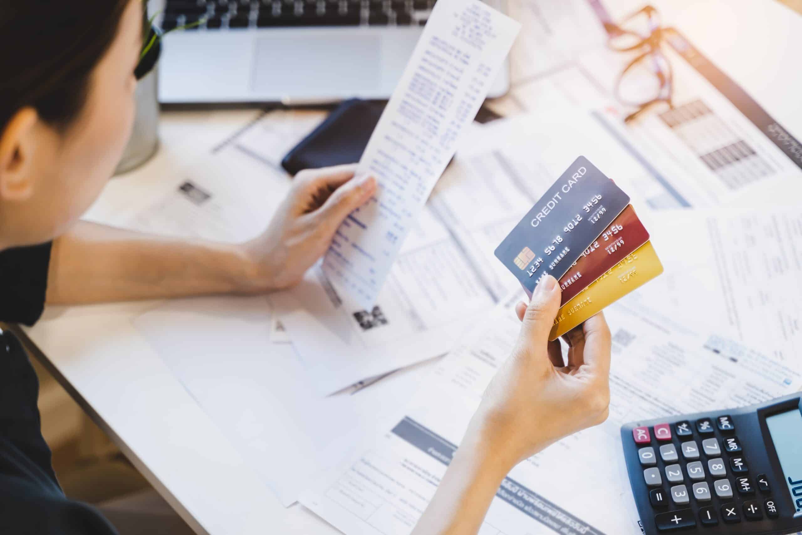 A person looks at a long receipt while holding two credit cards at a cluttered desk with bills, papers, laptop, and calculator.