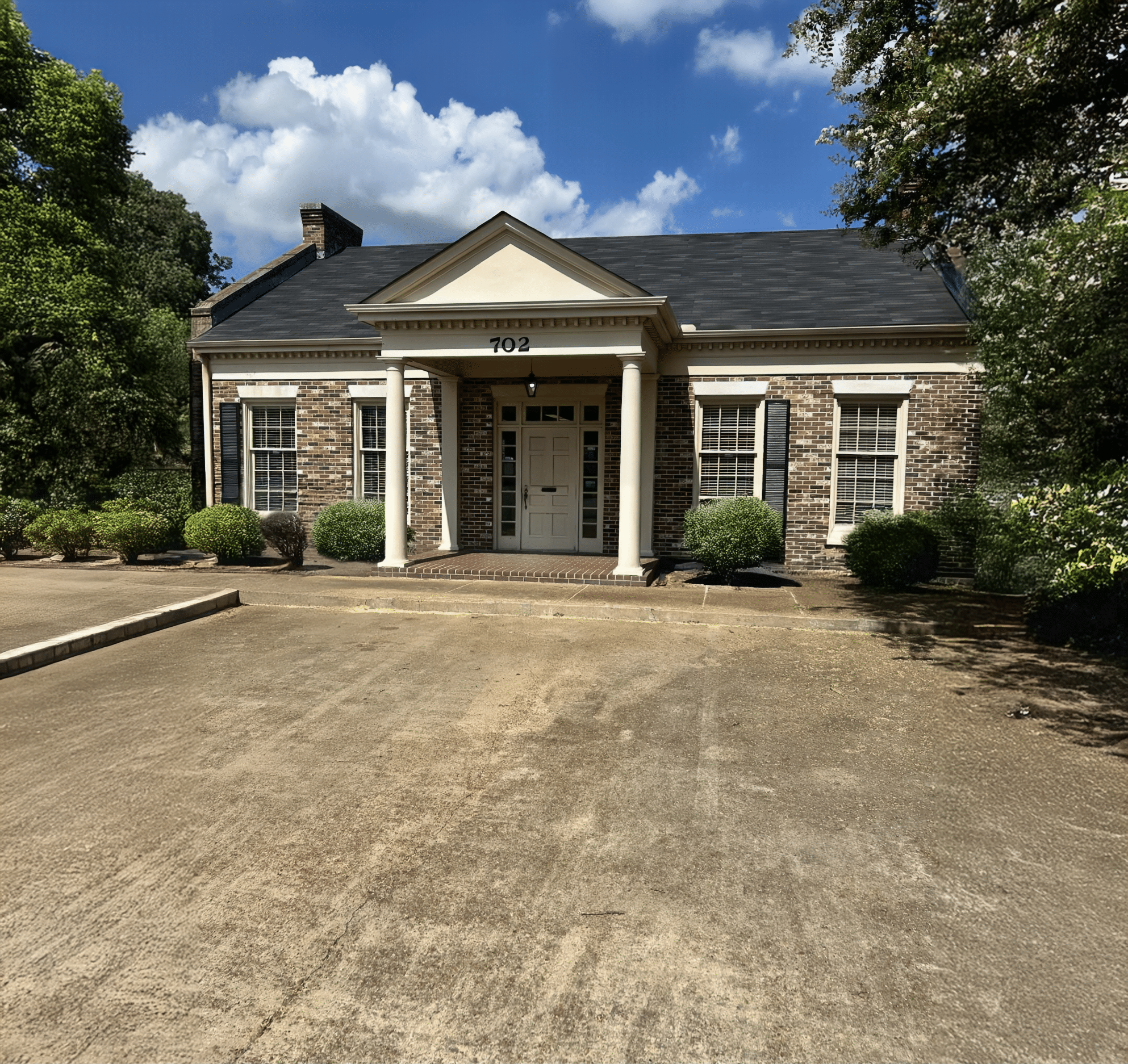 Small brick building with white columns, a black shingle roof, and 702 above the door, set amid bushes and trees under a cloudy sky.